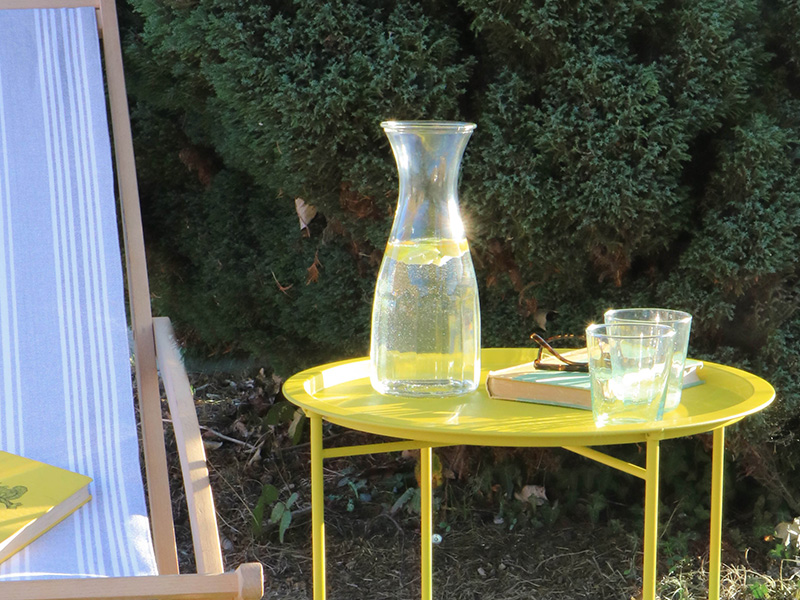 Rive Droite Outdoor Bistro Tray Table in Dandelion. Laid with a drinks carafe, glasses, book and sunglasses and next to a deckchair